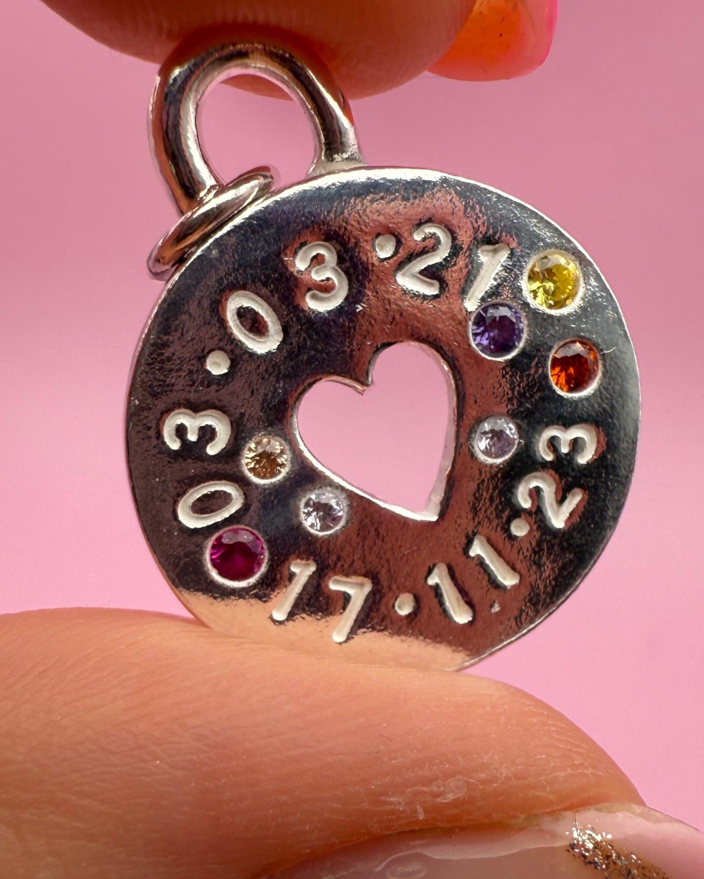 Close-up of a hand holding a circular metal charm with engraved numbers and colorful stones against a pink background.