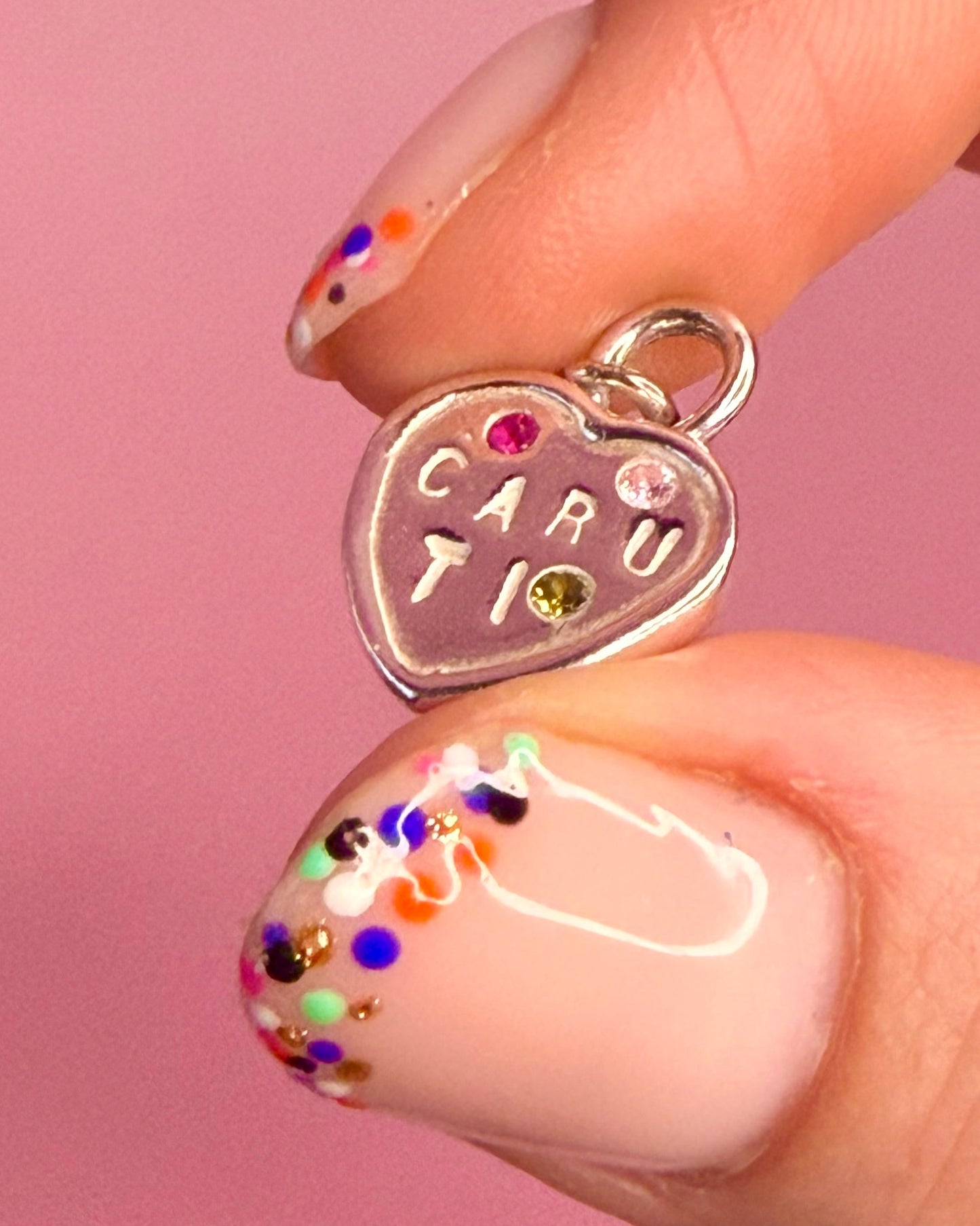 Heart-shaped silver pendant with 'caru ti' engraving and pink stones held between fingers against a pink background