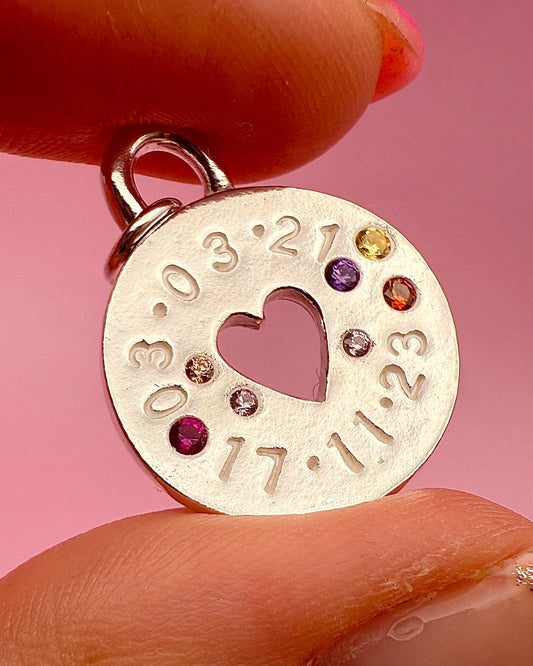 Silver charm with cut-out heart and engraved numbers held between fingers against a pink background