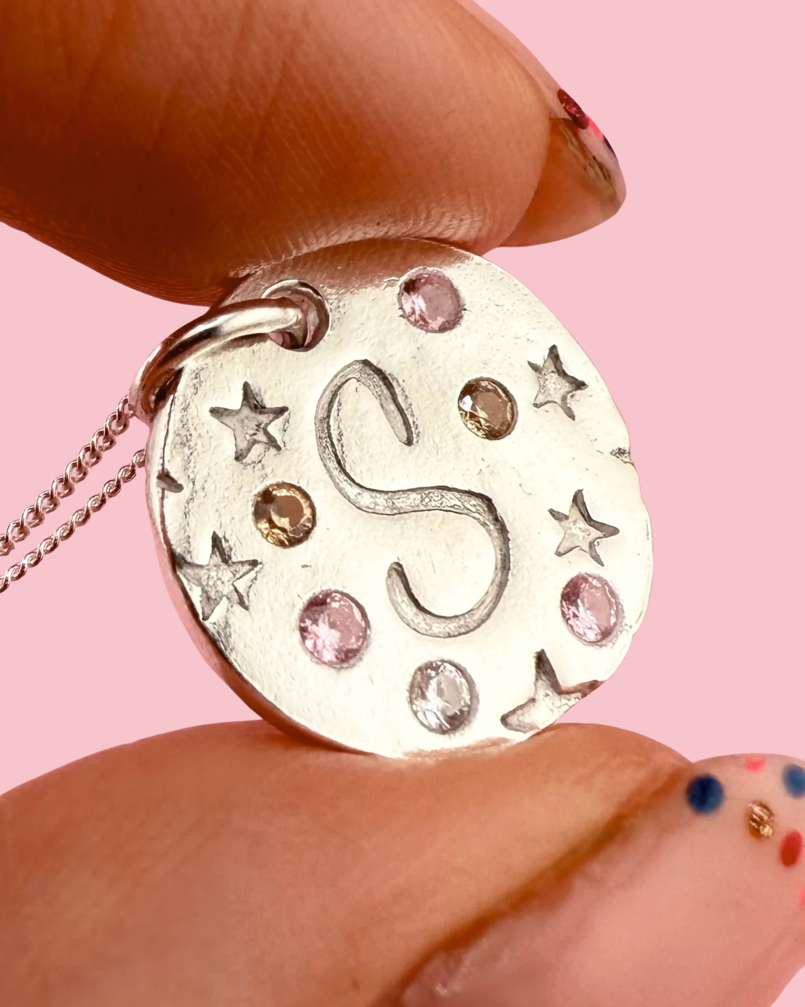 Circular Silver pendant with initial 'S' and decorative star elements held between fingers against a pink background