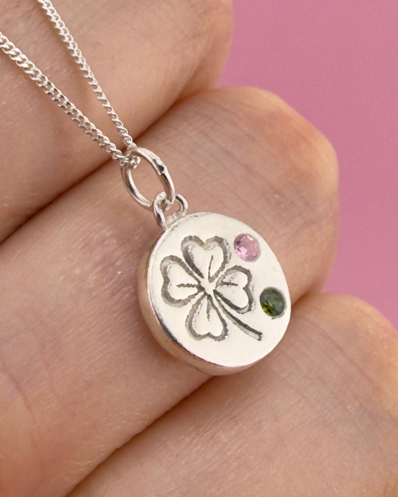 Silver necklace with a clover pendant held in a hand against a pink background