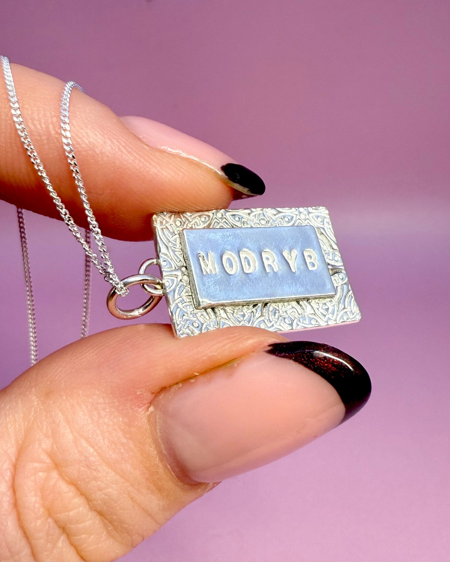 rectangular silver pendant with 'MODRYB' engraved on the front with celtic knot pattern on a pink background