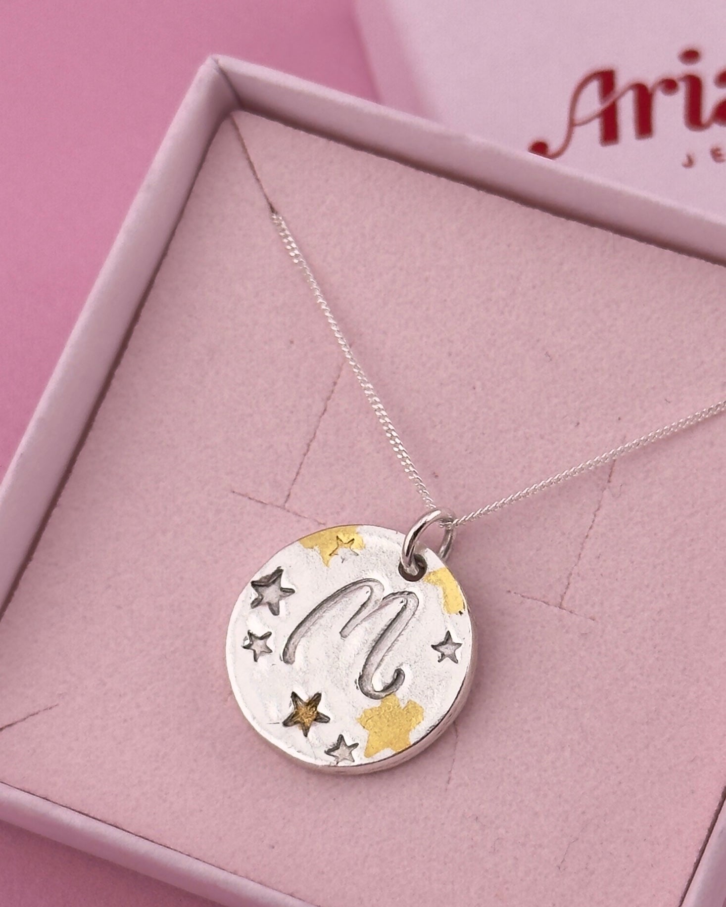 Silver necklace with a round pendant featuring stars and the letter 'M' on a pink box background.