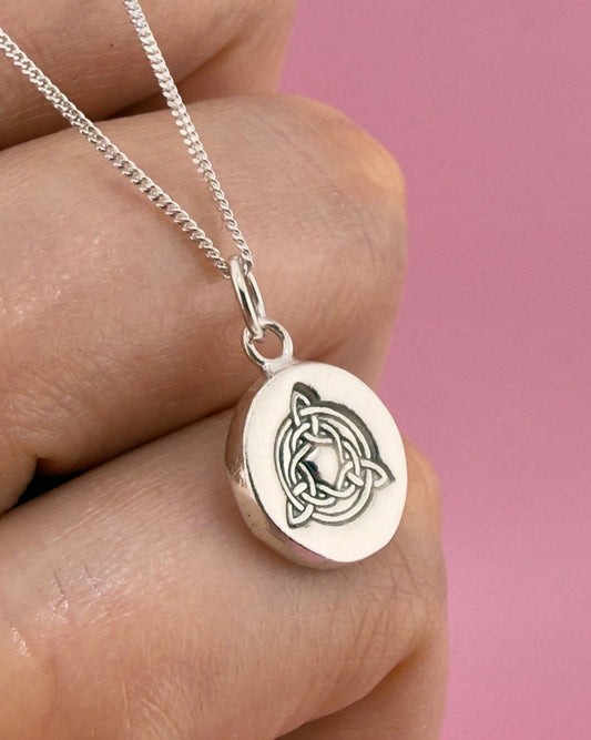 Silver necklace with a round pendant featuring a Celtic knot design, held between fingers against a pink background.