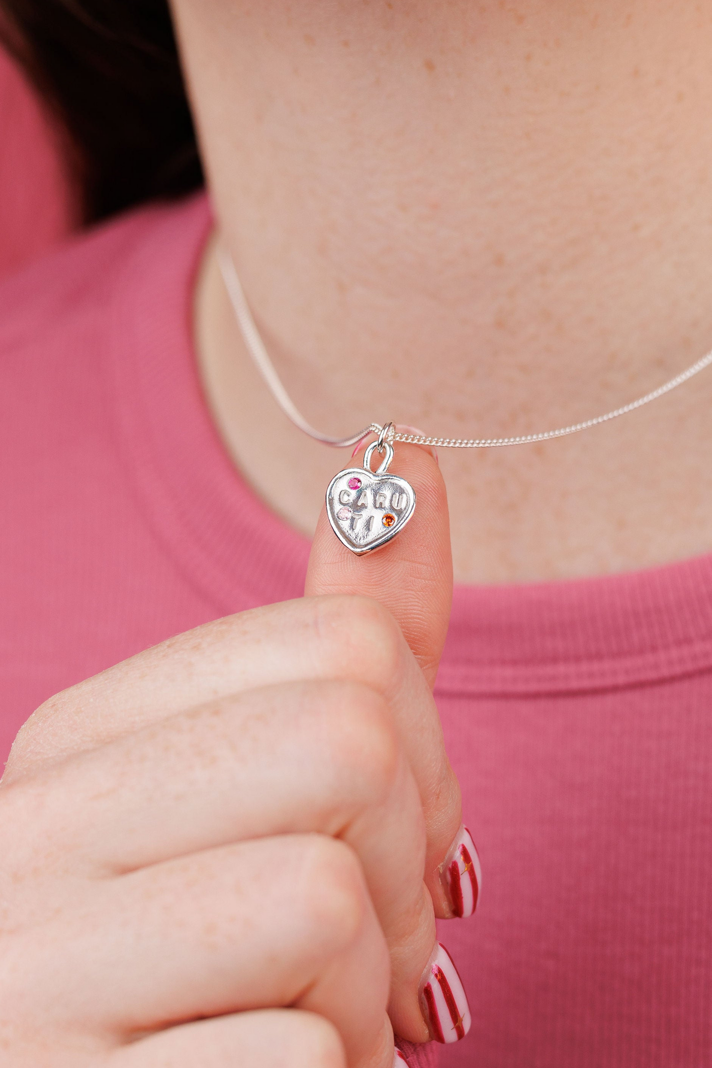 Person wearing a silver necklace with a heart-shaped pendant against a pink background