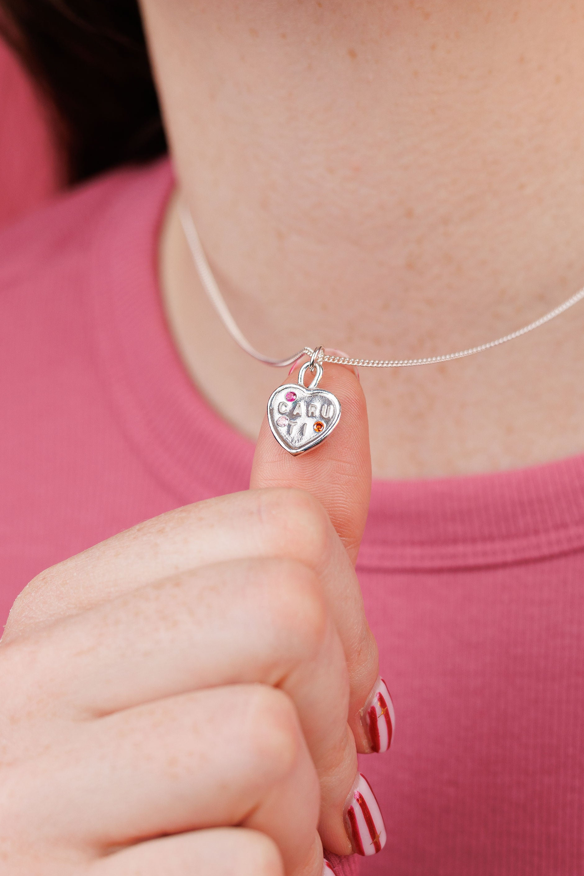 Person wearing a silver necklace with a heart-shaped pendant against a pink background
