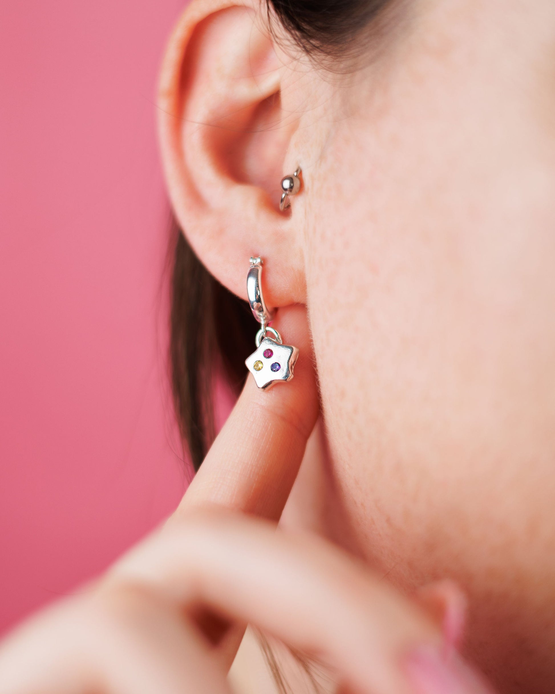 Close-up of a person wearing a silver earring with a small heart charm against a pink background