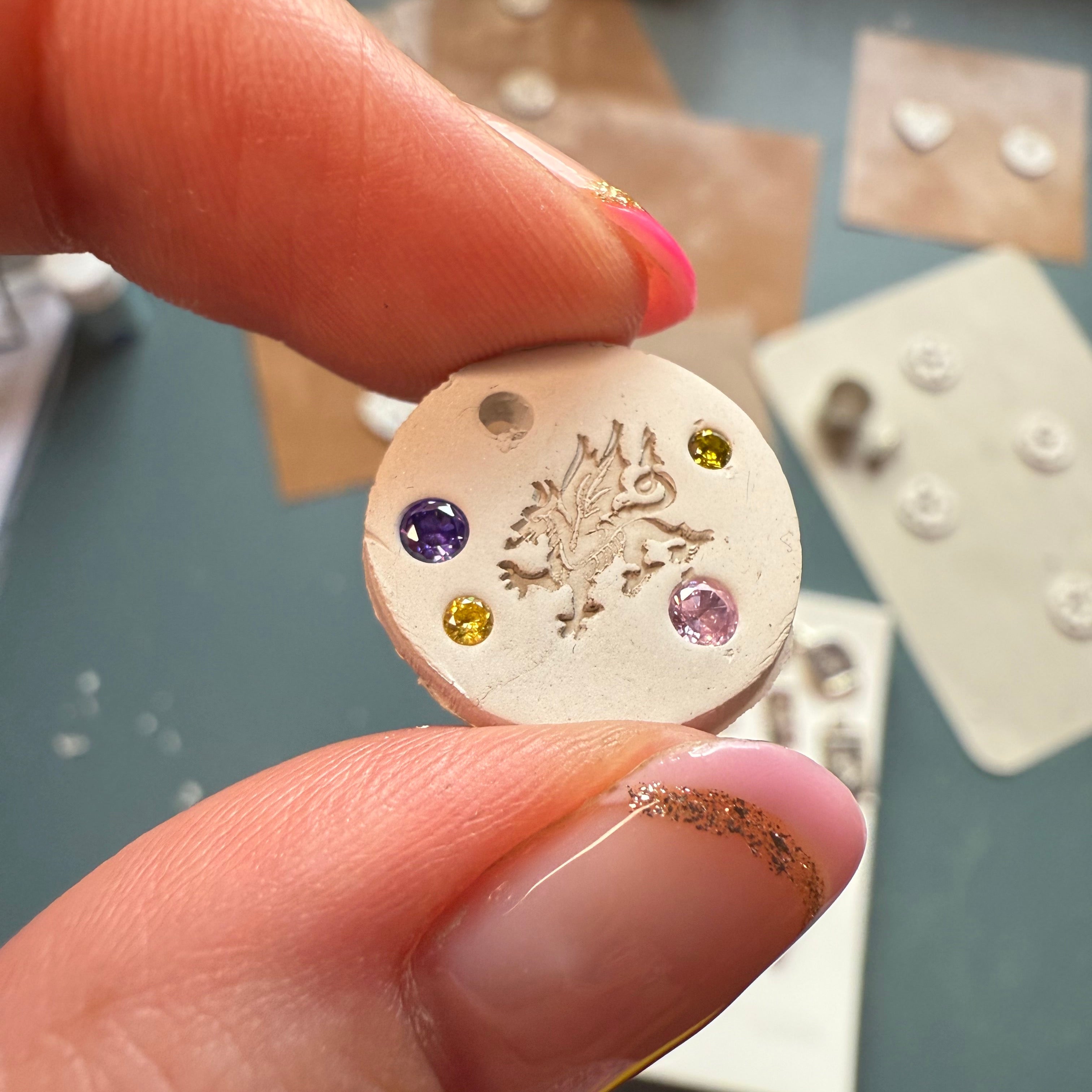 Close-up of a hand holding a dried silver clay charm with colourful gem stones against a blurred background.