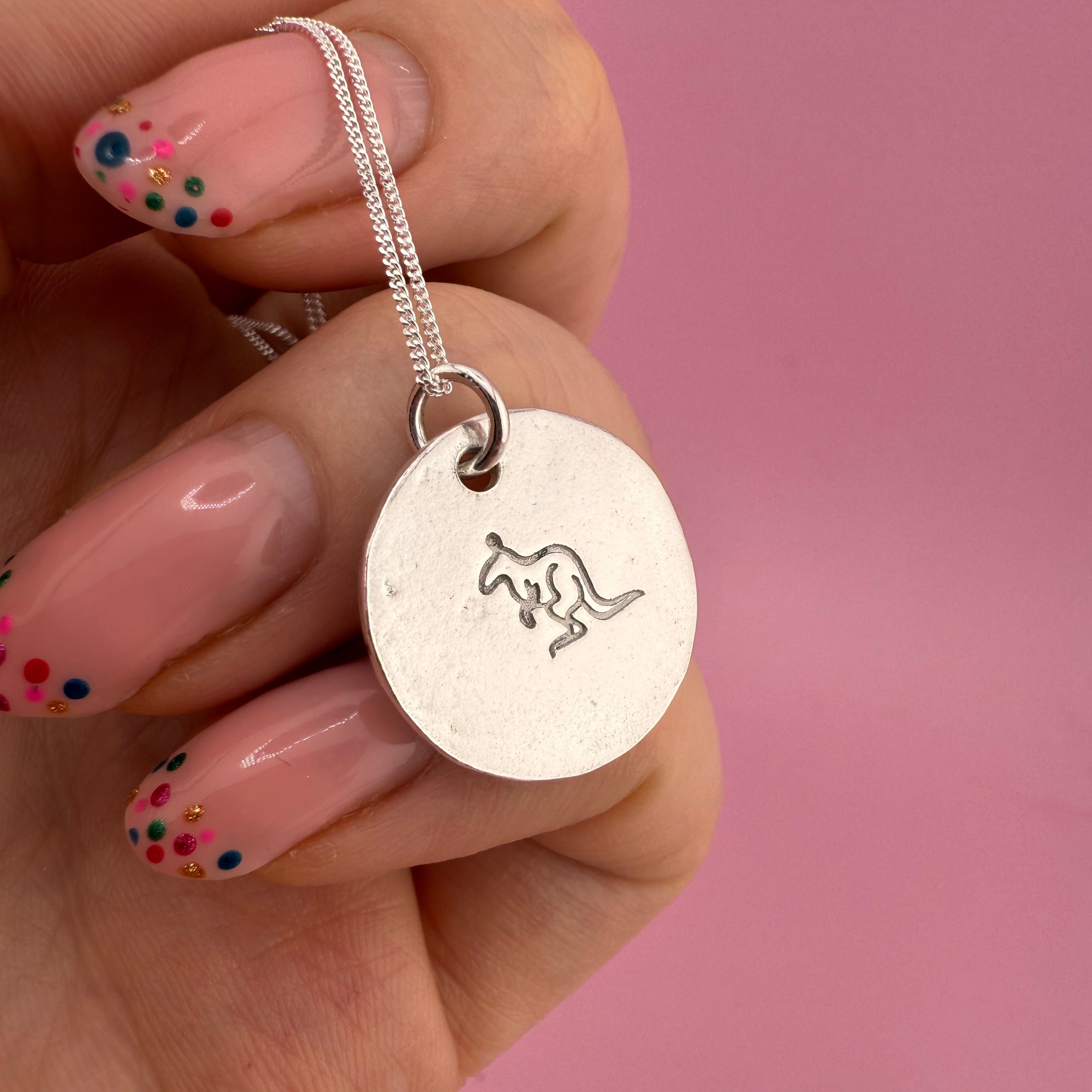 Hand holding a silver pendant with a kangaroo design against a pink background