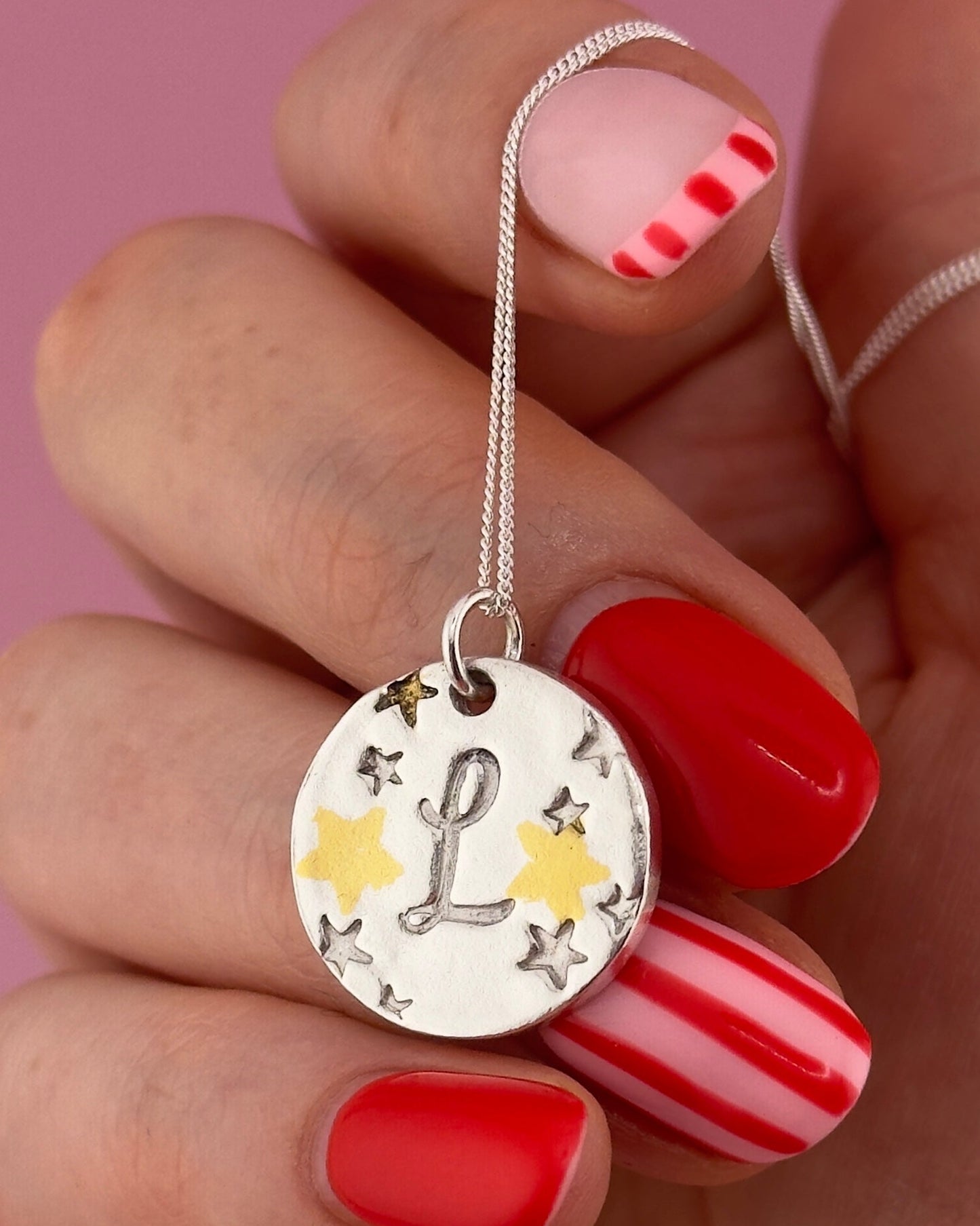 Hand holding a silver necklace with a pendant featuring the letter 'L' and stars against a pink background.